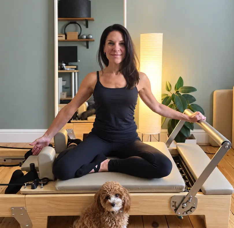 A teacher sitting cross-legged on a reformer with a small dog in front
